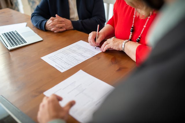 a-person-signing-a-document-at-a-table-with-others-with-a-laptop-and-paperwork-nearby