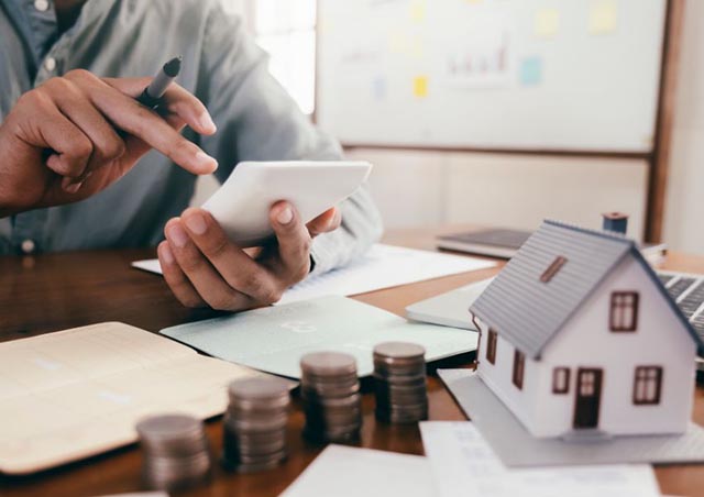 person using a calculator with coins and a house figurine in front of them on a table